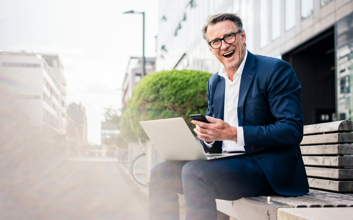 Man sitting on a bench with a laptop on his knees and a phone in his hand smiling